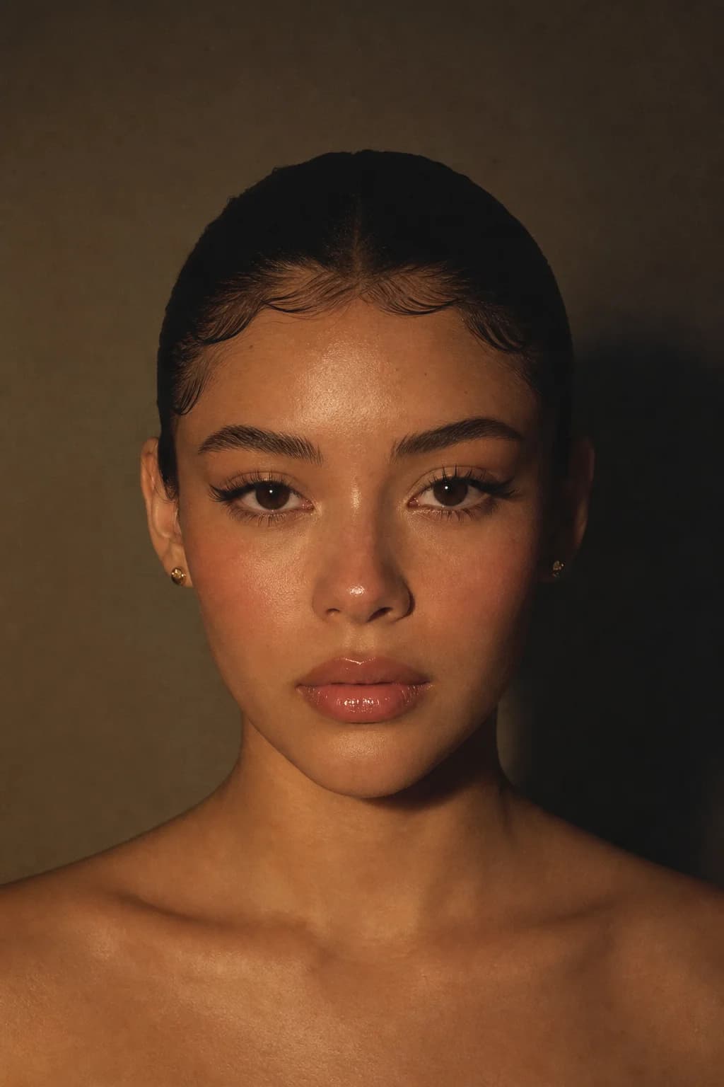 The same doe-eye makeup shot in low evening lamplight, head square to camera, lashes and glazed highlight catching the glow